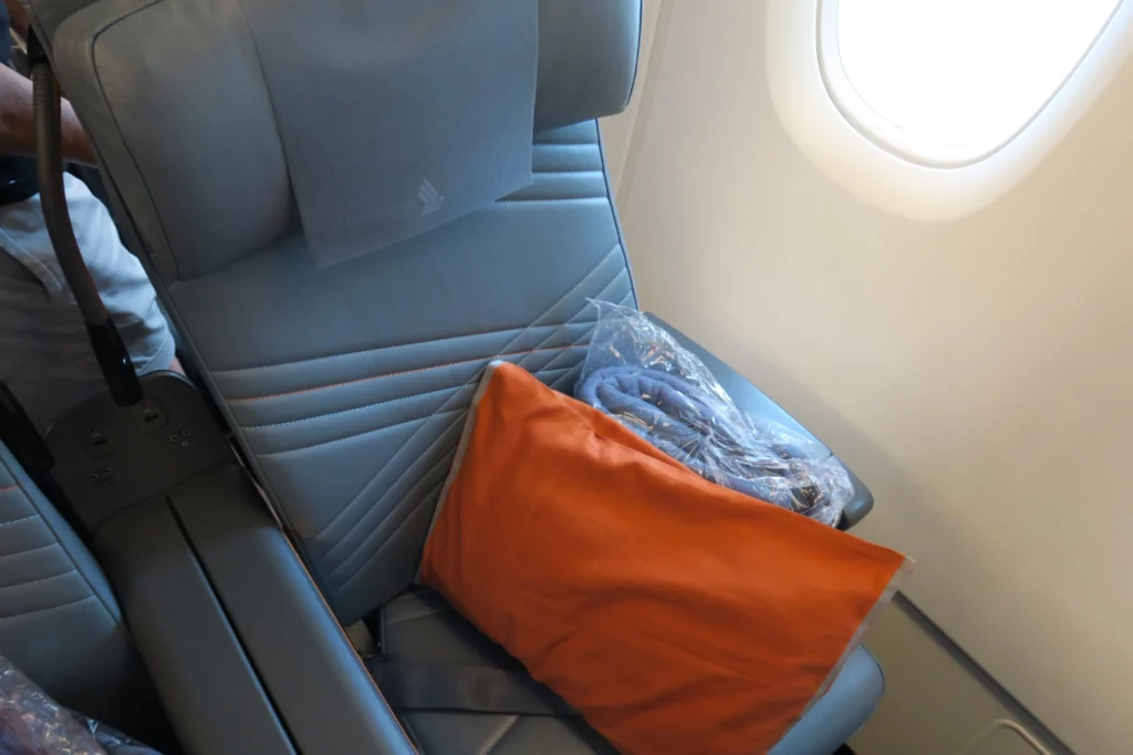 a seat with a pillow and an orange pillow next to an airplane window