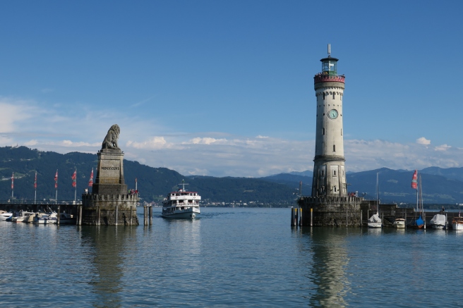 a lighthouse and a boat in the water with Lake Constance in the background